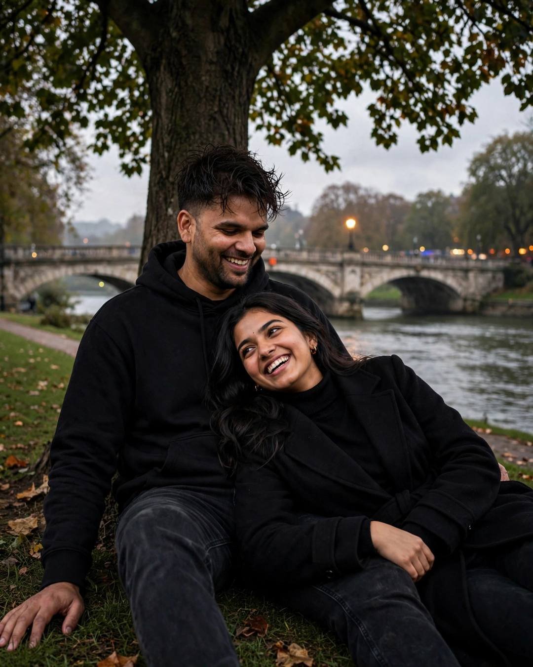 Couple laughing under a tree by a river with a stone bridge behind